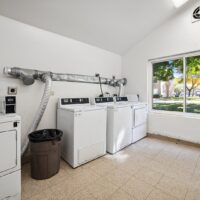 Small laundry room with three white washing machines, a trash can, tiled floor, and a large window showing trees and grass outside.