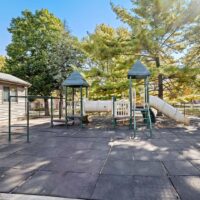 Outdoor playground with two slides, climbing structures, and rubber safety flooring, surrounded by trees and a fence on a sunny day.