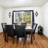 A dining room with a dark wooden table, six black chairs, hardwood floor, a window with sheer black curtains, and two decorative mirrors on white walls.