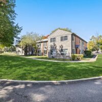 Two-story brick and siding apartment building with neatly trimmed lawn, trees, and a curved driveway under a clear blue sky.