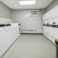 A small laundry room with multiple white washing machines and dryers, a wall-mounted air conditioner, and a fluorescent ceiling light.