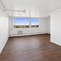 Empty room with white walls, large windows, brown wood flooring, a wall-mounted air conditioning unit, and a view of greenery and water outside.