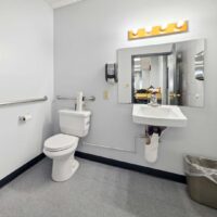 A public restroom with a toilet, wall-mounted sink, mirror, soap dispenser, grab bar, and trash can. Some cleaning supplies are visible in the mirror's reflection.