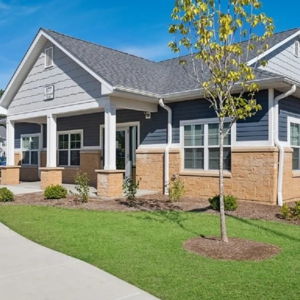 Single-story building with gray siding, tan brick lower walls, white trim, a covered entrance, and landscaped lawn on a sunny day.