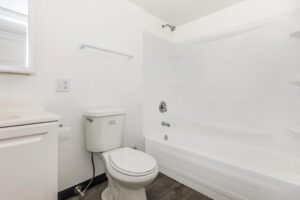 A clean, white bathroom features a toilet, bathtub with shower, built-in shelves, vanity with sink, and wood-look flooring.