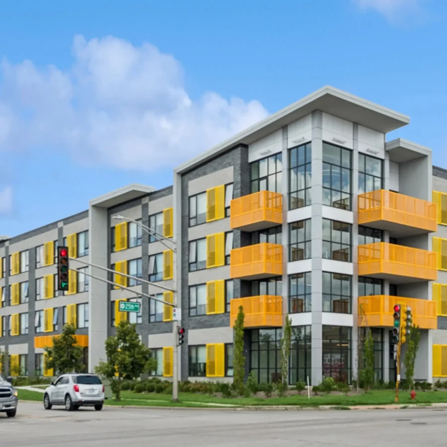 A modern four-story apartment building with large windows and bright yellow balconies and shutters, located at a street intersection with traffic lights and cars.