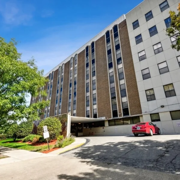 A multi-story building with a brick and concrete facade, a red car parked near the entrance, and trees and landscaping along the driveway.