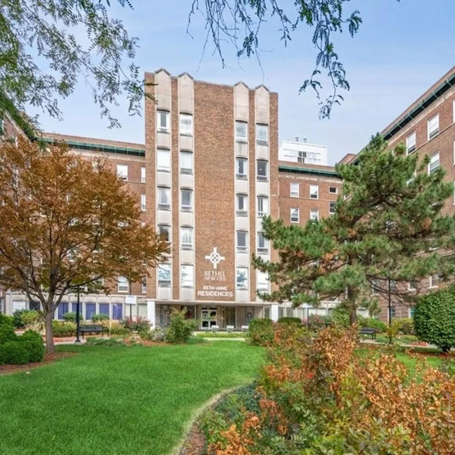 A multi-story brick residence hall with white windows and a central entrance sign, surrounded by green lawns, trees, and landscaped shrubs.