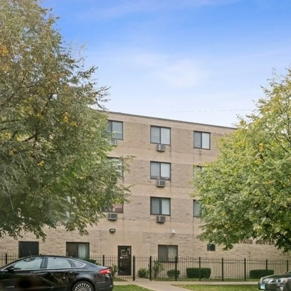 A three-story beige apartment building with multiple windows and window air conditioning units, partially obscured by trees, with parked cars and a fence in front.
