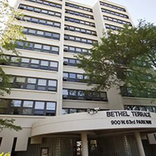 Multi-story building with white exterior and rows of windows, labeled "Bethel Terrace, 900 W. 63rd Parkway" above the entrance, with trees partially visible in the foreground.