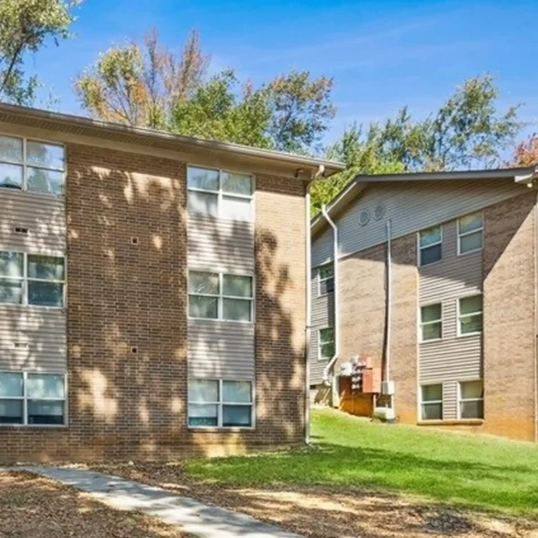 Two three-story apartment buildings with brick exteriors are surrounded by grass and trees under a clear blue sky.