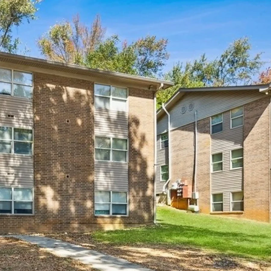 Two three-story apartment buildings with brick exteriors are surrounded by grass and trees under a clear blue sky.