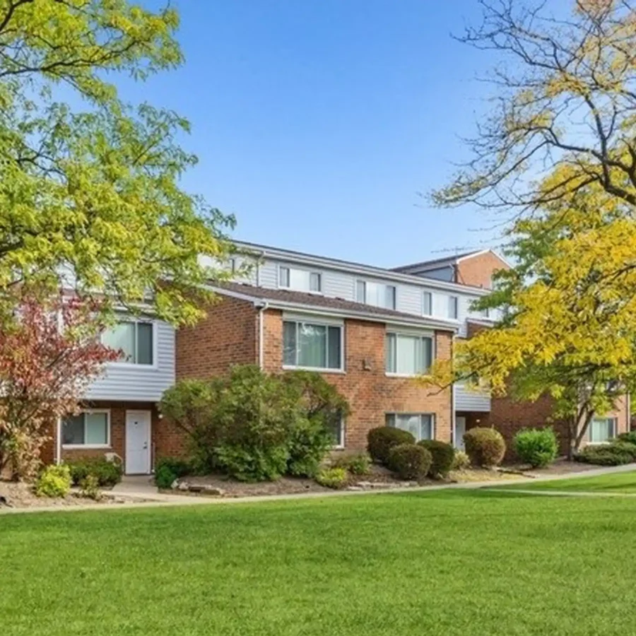 A two-story brick apartment building with large windows, surrounded by green lawns, trees, and shrubs under a clear blue sky.