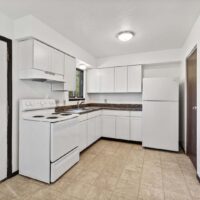 A small, empty kitchen with white cabinets, a white stove, a white refrigerator, a sink under a window, and beige tiled flooring.