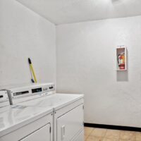 A small laundry room with two white machines, a broom resting on one, and a fire extinguisher mounted on a white wall.