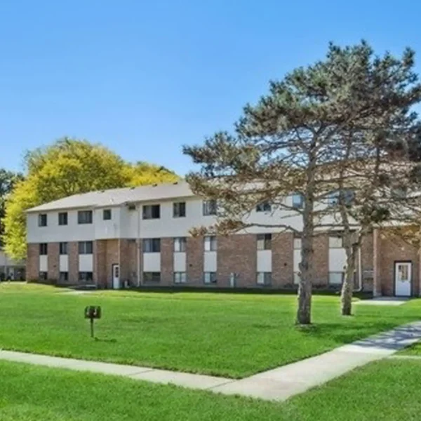 A three-story brick apartment building with several windows, surrounded by grass, trees, and concrete walkways on a clear day.