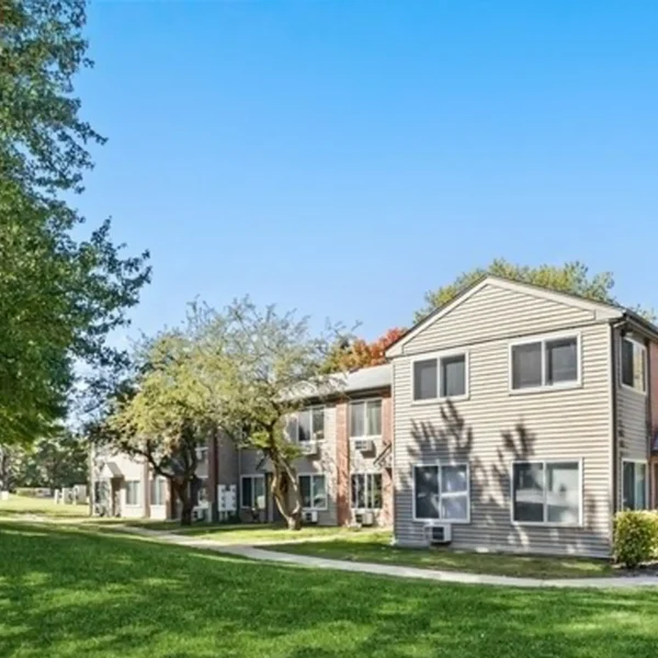Two-story apartment building with beige siding and multiple windows, surrounded by grass, trees, and a clear blue sky.