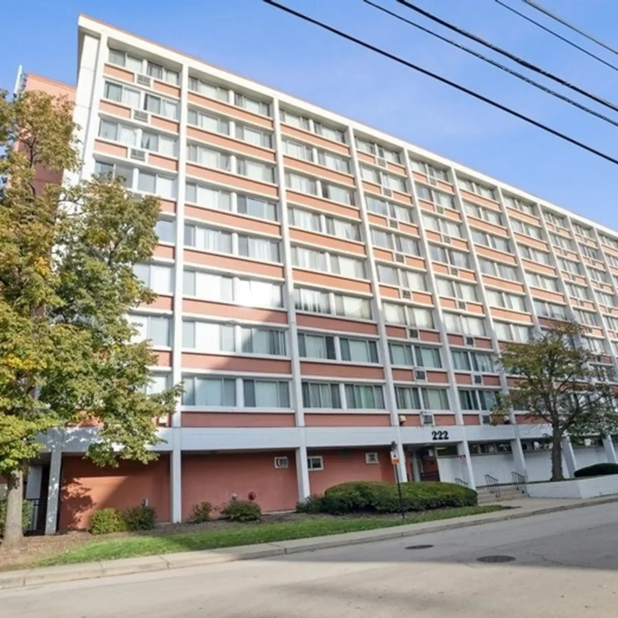 A multi-story apartment building with pink and white exterior panels, rows of windows, and the number 222 above the entrance, viewed from the street.