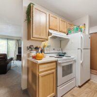 A compact kitchen with light wood cabinets, white appliances, a pumpkin on the counter, and boxed food on top of the refrigerator. The living room is visible in the background.
