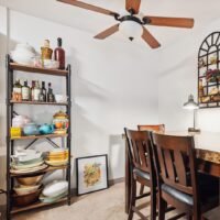 A metal shelf holds dishes, bowls, teapots, and bottles next to a dining table with four chairs; a framed picture rests on the floor against the wall.