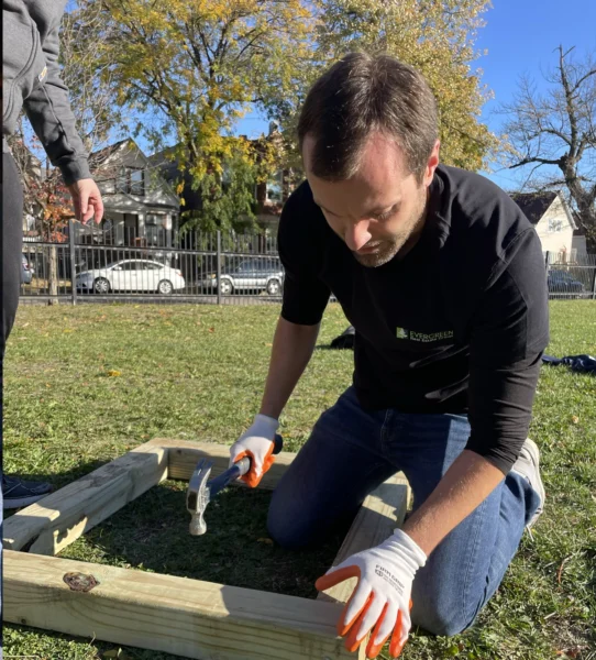 A man kneels on grass while hammering nails into a wooden frame. Another person stands nearby. Houses and trees are visible in the background.