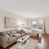 A tidy studio apartment with a gray sofa, white coffee table, bed with tufted headboard, large abstract wall art, and natural light from two windows.