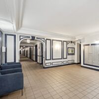 Apartment building lobby with blue armchairs, tiled floor, mailboxes on the right wall, and a hallway leading to apartments in the background.