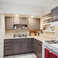 Modern kitchen with gray cabinets, white countertops, a gas stove with red towels, double sink, dish rack, open shelves with jars, and utensils hanging on a wall-mounted magnetic strip.