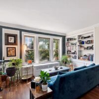 A bright living room with large windows, blue velvet sofa, wooden floors, indoor plants, a coffee table, and built-in bookshelves filled with books and framed photos.