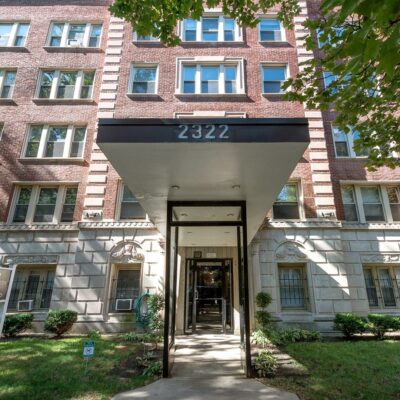 A brick apartment building with the number 2322 above the entrance canopy, surrounded by trimmed bushes and a small lawn.