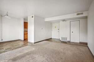 Unfurnished apartment interior with beige carpet, tile flooring near the entryway, white walls, two closets, and a small view of a kitchen with wooden cabinets.