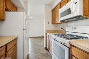 Galley kitchen with wood cabinets, white appliances including a refrigerator, microwave, and gas stove, and beige countertops; tile flooring extends to a dining area.