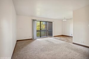 Empty living room with beige carpet, white walls, sliding glass door to a balcony, and dining area with tiled floor and a hanging light fixture.