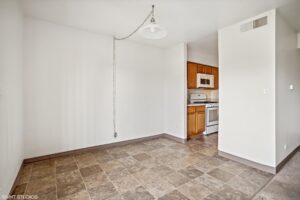 Empty room with tiled floor and hanging light fixture, adjacent to a small kitchen area with wood cabinets, a microwave, and a stove.