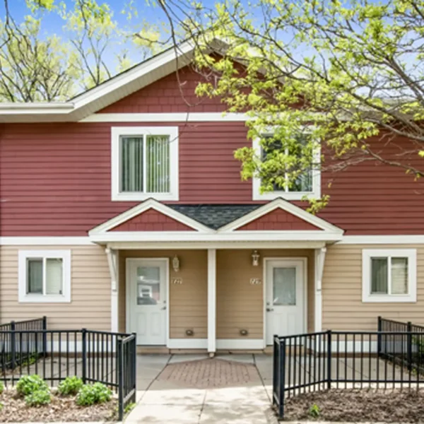 A two-story duplex with red and beige siding, white trim, two separate front doors, and black metal railings along the walkway.