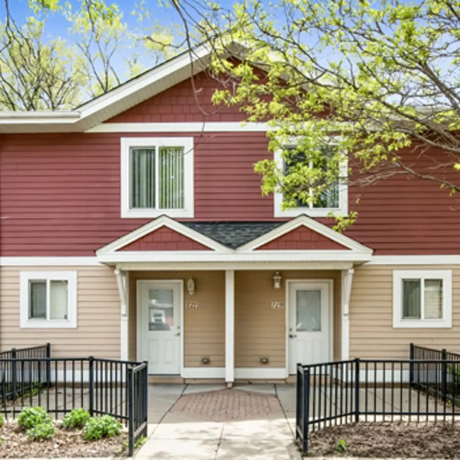 A two-story duplex with red and beige siding, white trim, two separate front doors, and black metal railings along the walkway.