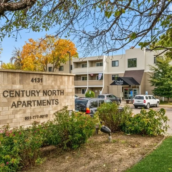 A beige sign reading "Century North Apartments" stands in front of a three-story apartment building with a parking lot and several cars.