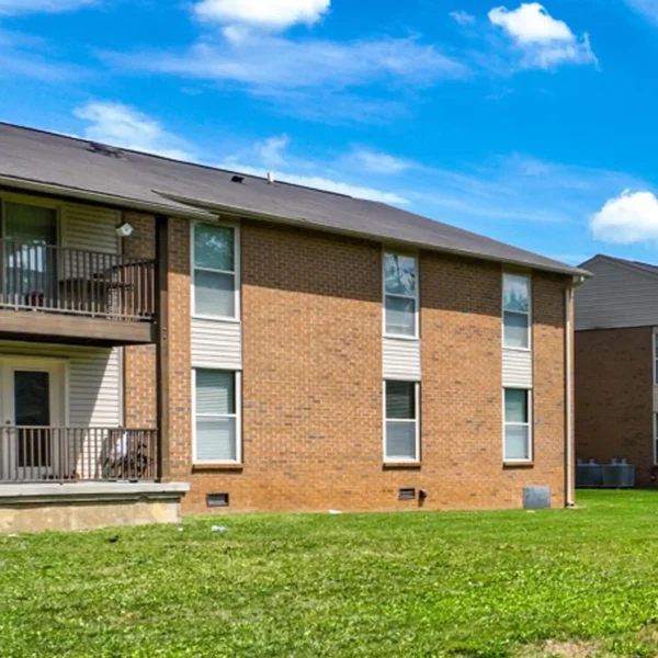 Two-story brick apartment building with balconies and white trim, situated on a grassy lawn under a blue sky with scattered clouds.