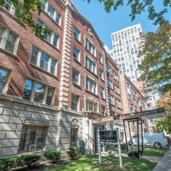 A multi-story brick apartment building with large windows and a "Now Leasing" sign out front, trees and a white van nearby, and a tall building visible in the background.