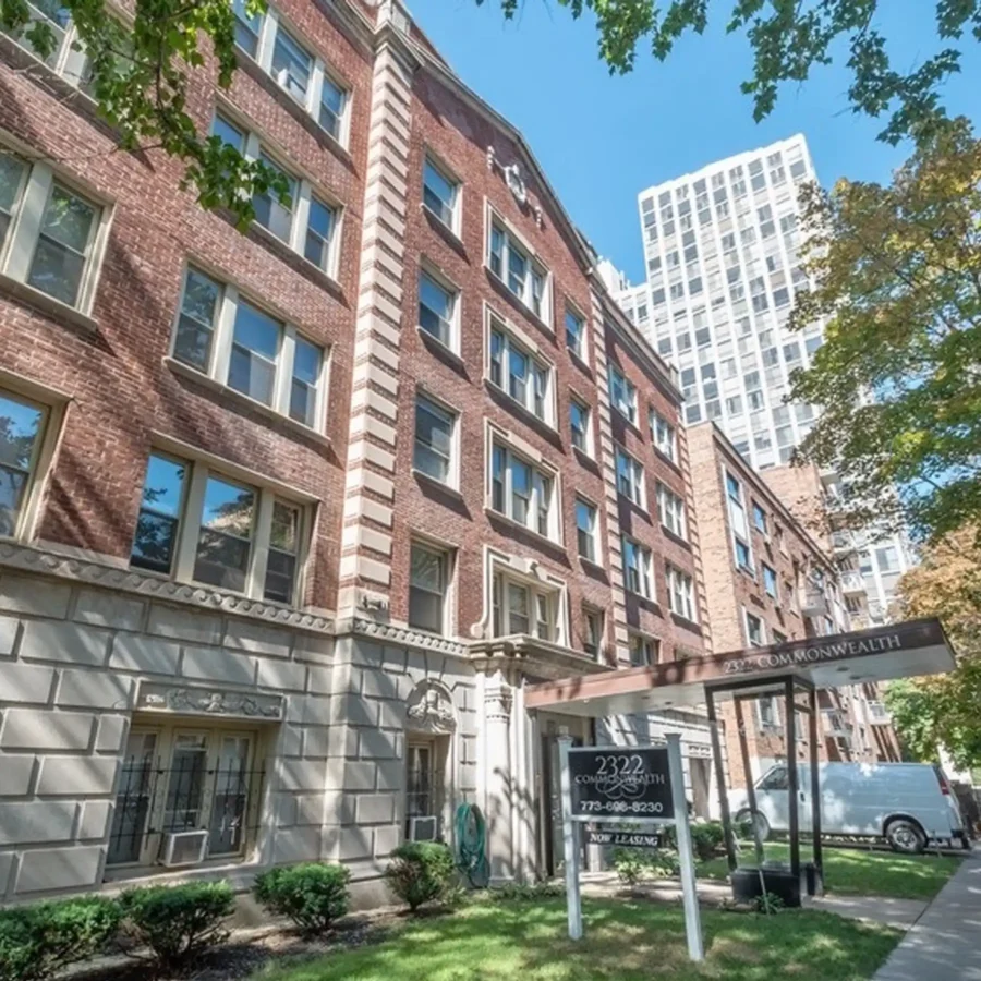 A multi-story brick apartment building with large windows and a "Now Leasing" sign out front, trees and a white van nearby, and a tall building visible in the background.