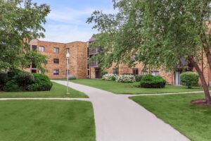 A courtyard with green lawns, trees, and bushes surrounded by brick apartment buildings and concrete walkways.