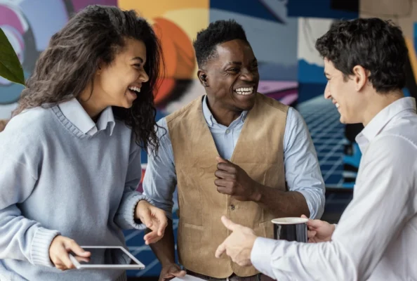 Three people are standing and talking together indoors; one holds a tablet, another holds a cup, and they are all smiling and laughing.