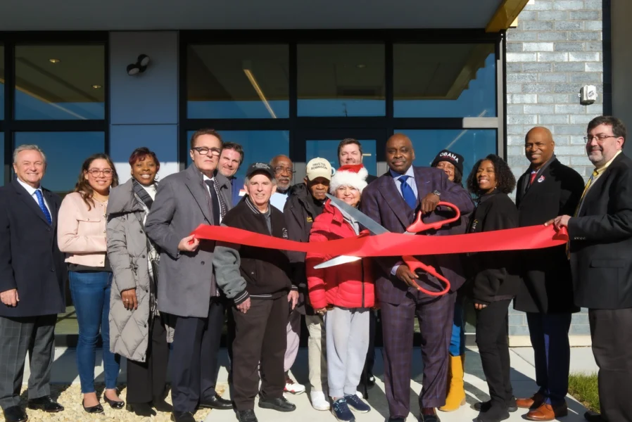 A group of people stands outside a building as two individuals cut a large red ribbon with oversized scissors during a ribbon-cutting ceremony.