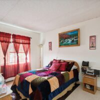 A bedroom with a colorful patchwork quilt on the bed, red curtains, wall art above the bed, and a small stereo on a nightstand. Natural light enters through the window.