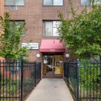 Entrance to a brick apartment building with a red awning labeled "3222," black metal fence, and trees on either side of a concrete walkway.