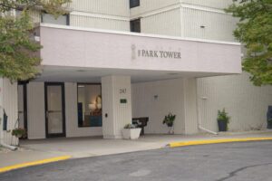 A beige building entrance with a sign reading "Park Tower" above the canopy. The address "247" is displayed near the door, with plants and trees around the entrance.