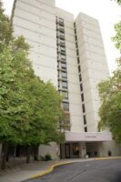 A tall, light-colored apartment or office building with multiple floors, partially obscured by green trees, and an entrance canopy labeled "FINAL TOWER.