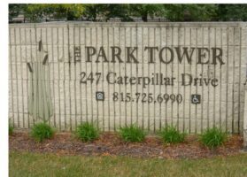 A stone sign displays "The Park Tower, 247 Caterpillar Drive, 815-725-6990" with icons for phone and wheelchair accessibility, and green plants at the base.