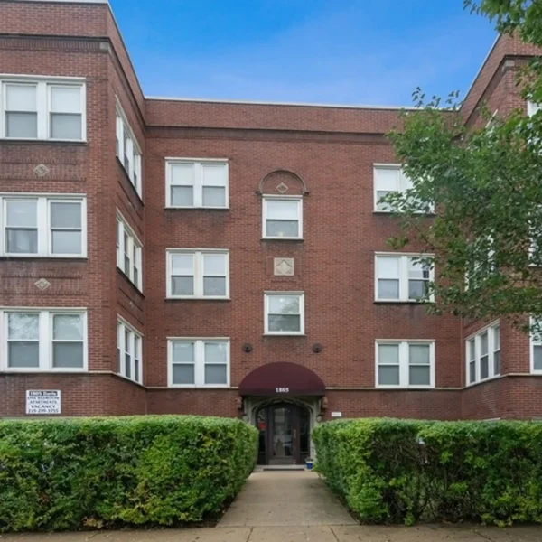 A three-story red brick apartment building with white-framed windows, a maroon awning over the entrance, and hedges lining the front walkway.