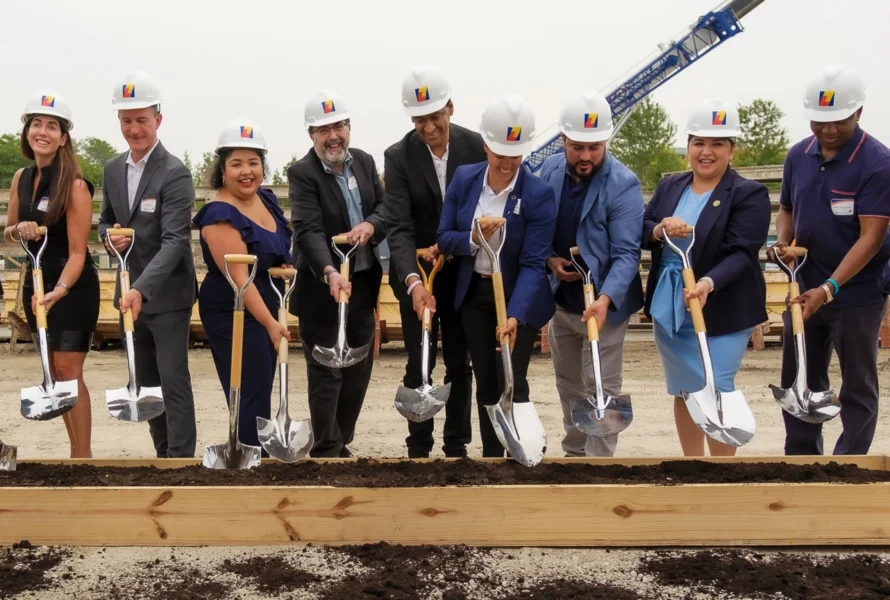 A group of nine people in hard hats and business attire participate in a groundbreaking ceremony, holding shovels and standing in front of a wooden frame filled with soil.